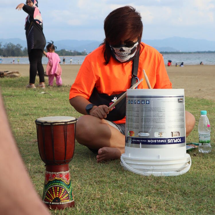 Gin (in skull mask) drumming a repurposed paint bucket, in front of them there is a mini djembe.